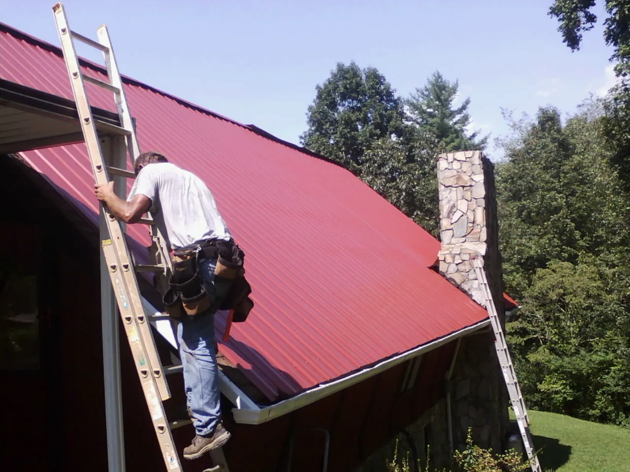 Roofer on a ladder beside a red metal roof and stone chimney — Soffit & Fascia Repair by Roofing Brothers in Ellensburg